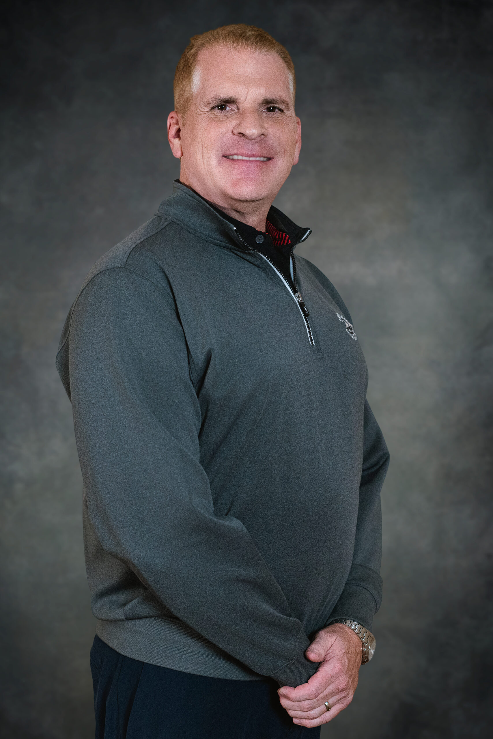 Pastor Bob Bryant stands with short hair groomed, wearing a gray half-zipped pullover, and stands against a dark gray mottled background. He is facing slightly to the right with a relaxed smile and his hands are crossed in front of his body.