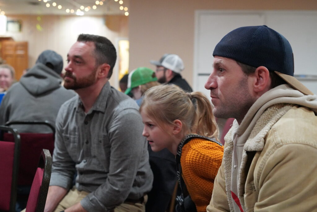 A group of people sits attentively indoors, focused on the church's service ahead. A man in a gray shirt and another in a tan jacket with a black cap sit on either side of a young girl in an orange sweater. The background is softly lit with fairy lights.