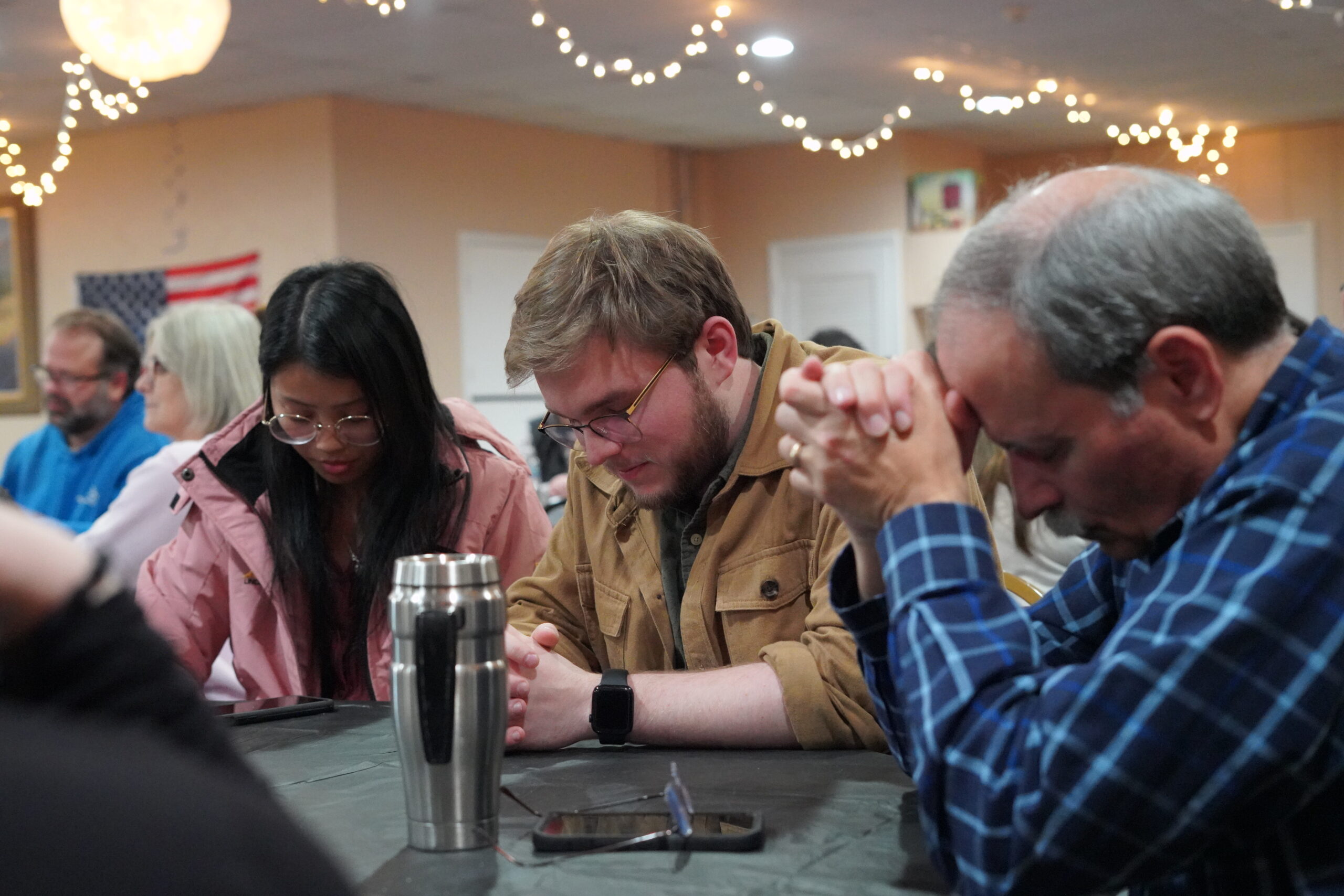 A group of people sitting at a table, heads bowed in prayer. The room is decorated with string lights and an American flag is partially visible in the background. A stainless steel tumbler and phones are on the table.