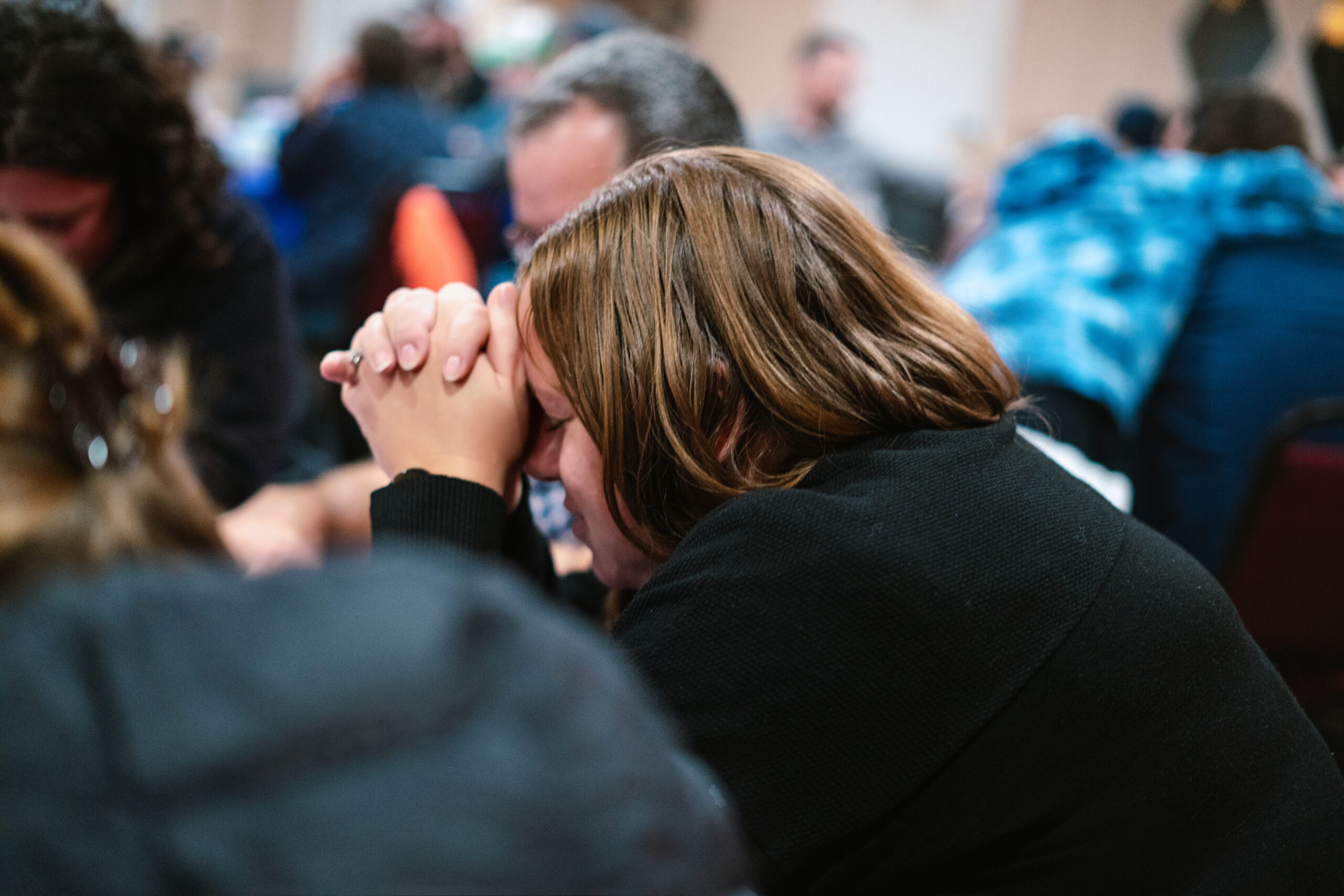 A person with brown hair rests their head on clasped hands, appearing deep in prayer. They are seated at a table with others in the background as a group setting. The lighting is soft and warm.