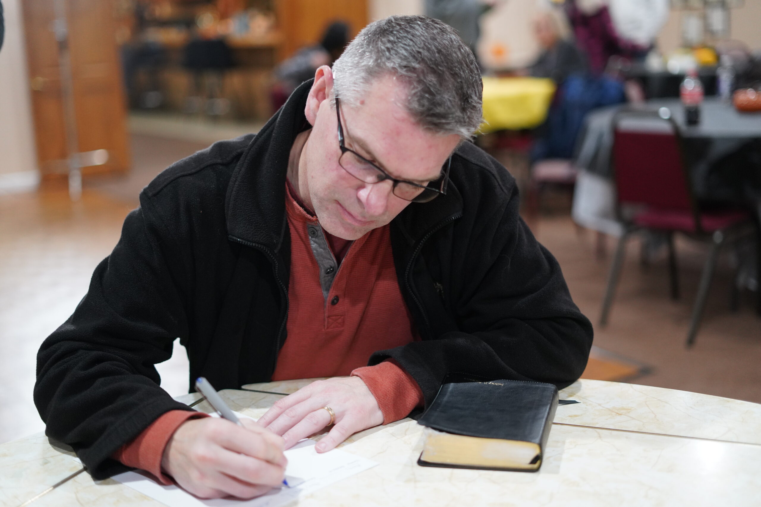 A man wearing glasses, sitting at a table, writing on paper with a pen. A Bible is placed beside him. The background shows a blurred indoor setting with more chairs, tables, and figures.